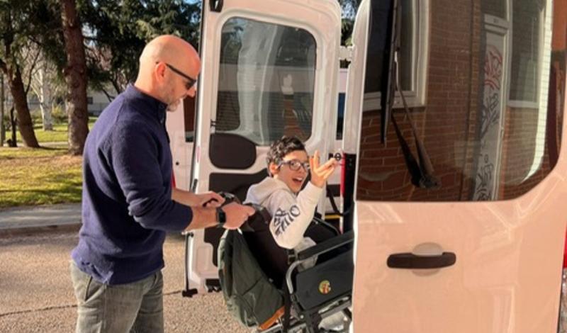 Aparece un niño en su silla de ruedas eléctrica subiendo a la plataforma del vehículo adaptado ayudado por personal de AENILCE