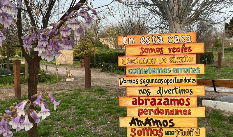 Imagen de un paseo arbolado con flores rosáceas, delante de los arboles en el margen derecho de la imagen hay panel informativo descriptivo del lugar al que llegamos y dice :  En esta casa somos reales decimos lo siento, cometemos errores, damos segundas oportunidades, nos divertimos, abrazamos, perdonamos, amamos,somos una familia.