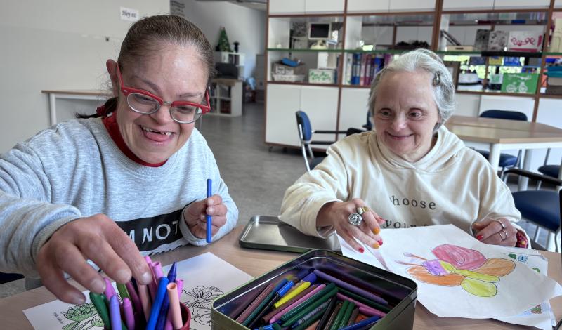 Dos personas mayores con discapacidad intelectual, sonrien mientras pintan con ceras en unos folios sobre una mesa. En el centro una caja metálica y un cubilete lleno de pinturas de cera.
