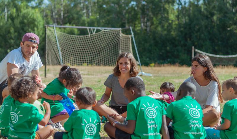 9 niños y niñas de espaldas con camisetas verdes sentados en círculo en el campo jugando a algo con 1 monitor y dos monitoras (una sonríe la otra les habla). Los niños y niñas tienen entre 2 y 7 años visten camisetas verdes de la Fundación Soñar despierto, dos de ellos son de raza negra y de al menos 3 de ellos se distingue su nombre escrito en la espalda (Luis, Ossas y Regi)