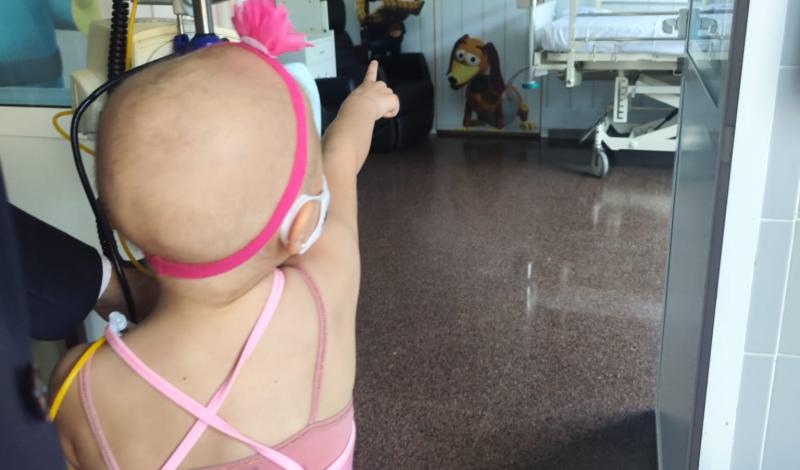 Niña pequeña afectada por cáncer, con ausencia de cabello, vestida con un tutú rosa y conectada a un equipo médico, señala hacia una habitación hospitalaria decorada con personajes de Toy Story, donde se observa una cama clínica al fondo.