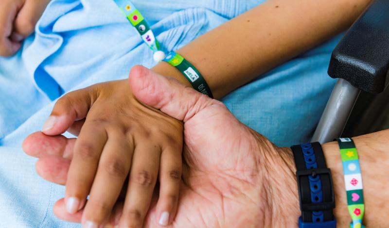 En la fotografía aparece una mano de una persona sosteniendo la mano de una segunda persona que está sentada en una silla. Ambas personas llevan una pulsera en la que se lee AdELA en las muñecas.