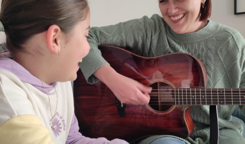 EN LA IMAGEN SE VEN DOS PERSONAS, UNA MUJER TOCANDO LA GUITARRA Y UNA NIÑA ESCUCHANDO LO QUE TOCA