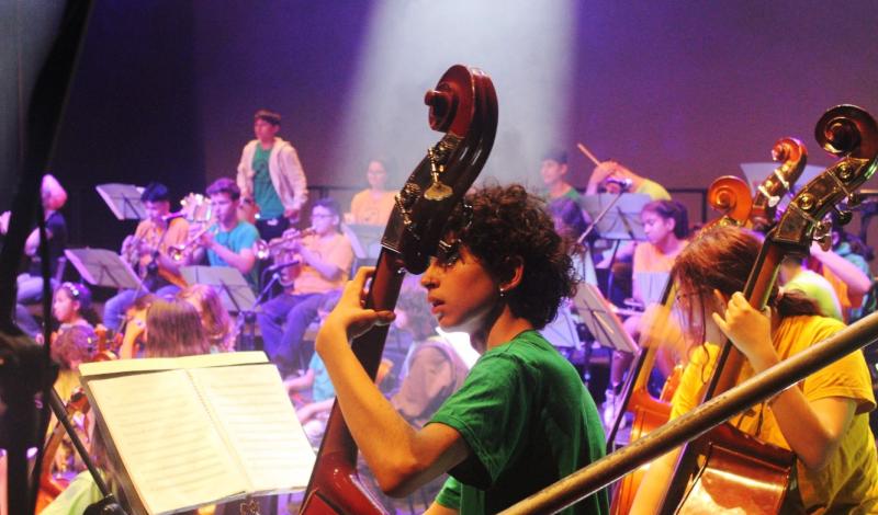 Grupo de adolescentes, jóvenes y niños/as que en conjunto forman la Orquesta DaLaNota tocando diferentes instrumentos (violines, trompetas, percusión...) junto a las diferentes partituras.