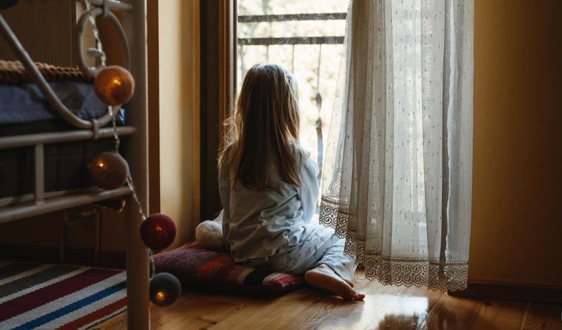 Es una niña pequeña, en pijama y descalza, sentada en el suelo de espaldas mirando por una ventana.