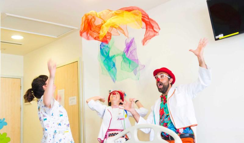 Aparecen tres personas en la imagen, dos payasos de hospital lanzando un pañuelo al aire de colores a una paciente ingresada.