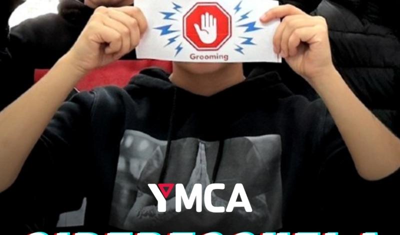 En la imagen aparecen varios menores en situación de vulnerabilidad social participando en una actividad de sensibilización. Cada uno sostiene una tarjeta rectangular con el símbolo de “Grooming”, representado por una mano levantada dentro de un hexágono rojo, acompañada de líneas azules que simulan una alerta. Se encuentran en un espacio interior iluminado, probablemente un aula o sala de actividades. En la parte inferior de la imagen se muestra el logotipo de YMCA, seguido del título  “CIBERESCUELA"