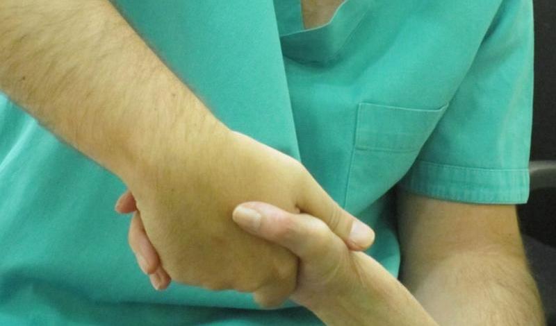 “Una persona con bata médica verde sostiene con una mano la mano de un paciente, en un gesto de apoyo" y cuidado