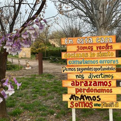 Imagen de un paseo arbolado con flores rosáceas, delante de los arboles en el margen derecho de la imagen hay panel informativo descriptivo del lugar al que llegamos y dice :  En esta casa somos reales decimos lo siento, cometemos errores, damos segundas oportunidades, nos divertimos, abrazamos, perdonamos, amamos,somos una familia.