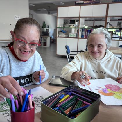 Dos personas mayores con discapacidad intelectual, sonrien mientras pintan con ceras en unos folios sobre una mesa. En el centro una caja metálica y un cubilete lleno de pinturas de cera.