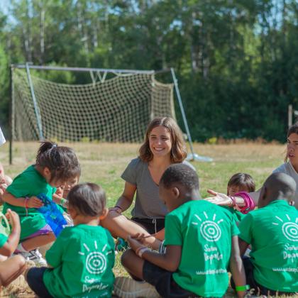 9 niños y niñas de espaldas con camisetas verdes sentados en círculo en el campo jugando a algo con 1 monitor y dos monitoras (una sonríe la otra les habla). Los niños y niñas tienen entre 2 y 7 años visten camisetas verdes de la Fundación Soñar despierto, dos de ellos son de raza negra y de al menos 3 de ellos se distingue su nombre escrito en la espalda (Luis, Ossas y Regi)