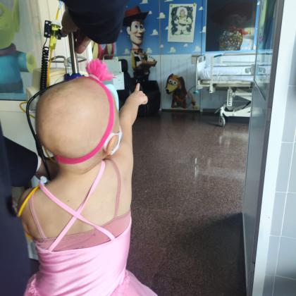 Niña pequeña afectada por cáncer, con ausencia de cabello, vestida con un tutú rosa y conectada a un equipo médico, señala hacia una habitación hospitalaria decorada con personajes de Toy Story, donde se observa una cama clínica al fondo.