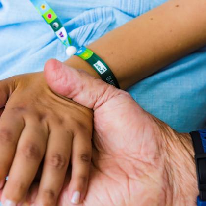 En la fotografía aparece una mano de una persona sosteniendo la mano de una segunda persona que está sentada en una silla. Ambas personas llevan una pulsera en la que se lee AdELA en las muñecas.