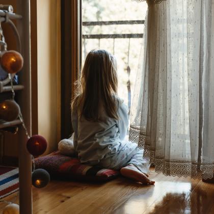 Es una niña pequeña, en pijama y descalza, sentada en el suelo de espaldas mirando por una ventana.