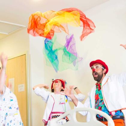Aparecen tres personas en la imagen, dos payasos de hospital lanzando un pañuelo al aire de colores a una paciente ingresada.