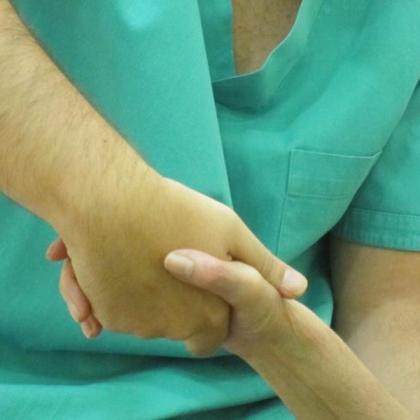 “Una persona con bata médica verde sostiene con una mano la mano de un paciente, en un gesto de apoyo" y cuidado